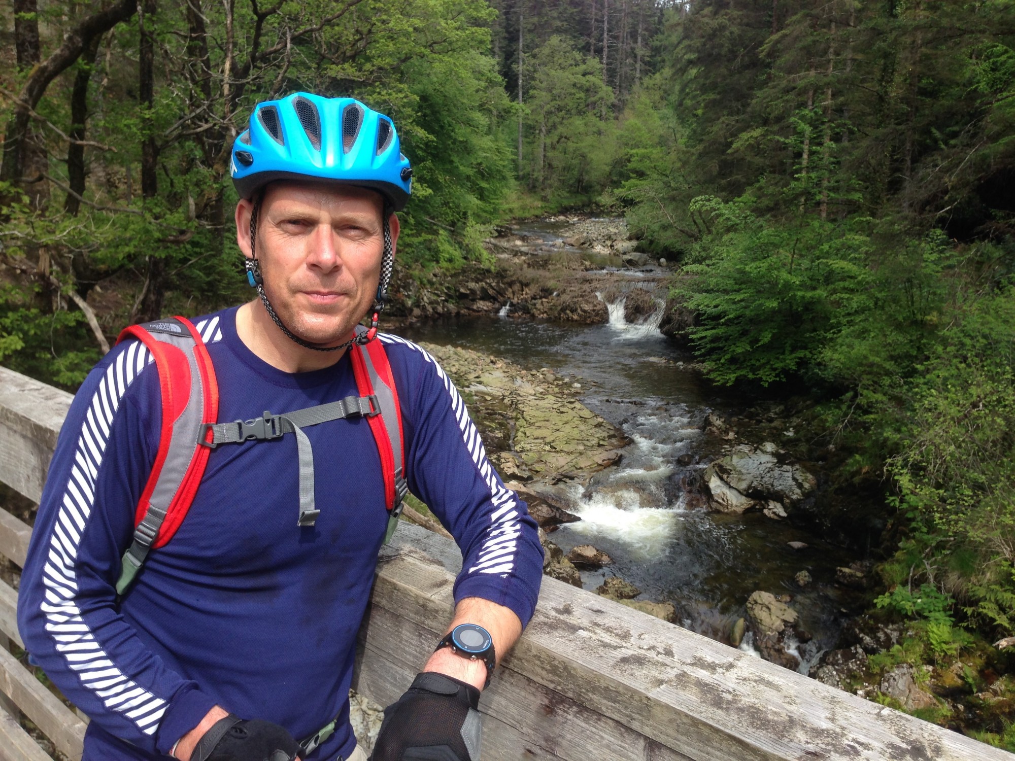 Carl stands in front of a waterfall, looking at the camera and wearing a cycling helmet and rucksack. 