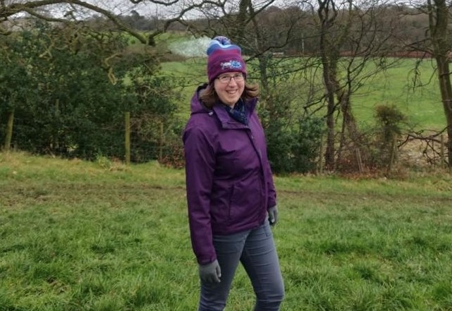 Helen wears a hat and smiles. She is standing in a filed with trees behind her.