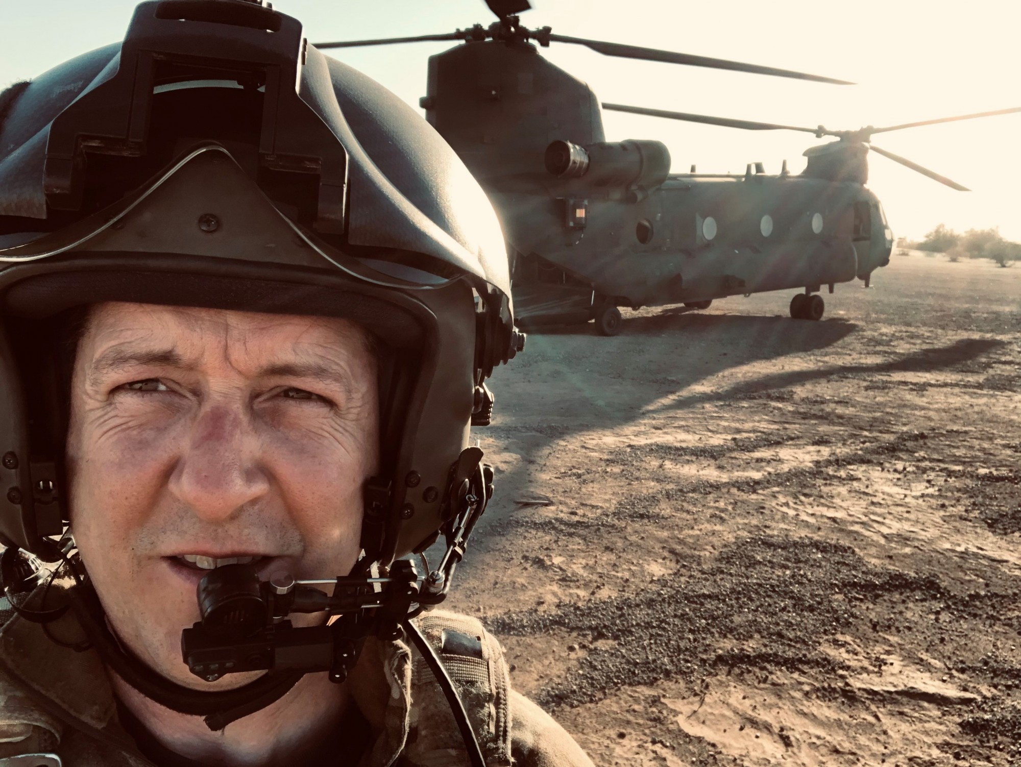 A close up of Ian Swift wearing a helmet with a Chinook helicopter behind him the desert